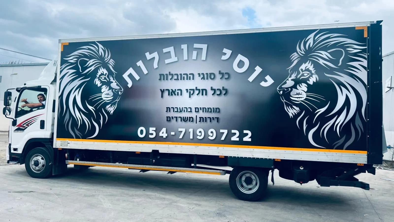 המשאית של יוסי הובלות מהצד, עם שני האריות, השם ומספר הטלפון, ויוסי בחלון הנהג מרים אגודל